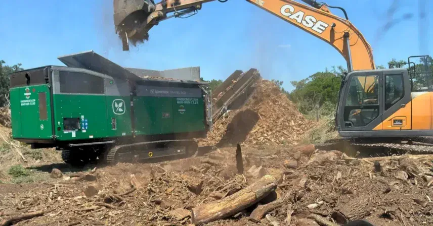 Excavator feeding wood debris into a large grinder during land clearing, with piles of mulch and tree remains scattered across the site.